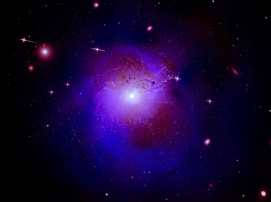 A bright white spot at the center of the image is surrounded by a blue and red halo. The blue represents X-ray emission and appears as a thick “0” around the bright center. The red halo is optical and radio light, which fills the middle of the X-ray emission. Dotted around the image are additional white spots, which are galaxies and foreground stars.
