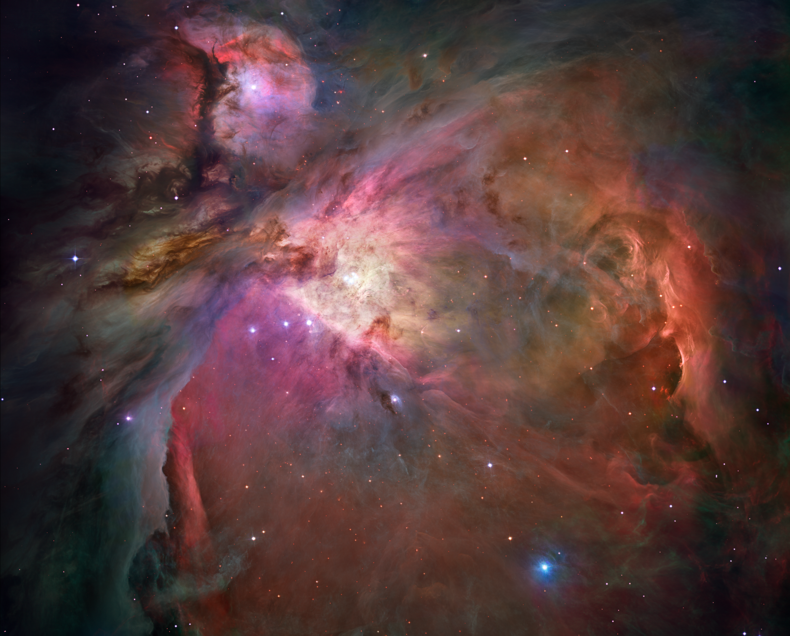 This Hubble image of the Orion Nebula offers a peek inside a cavern of roiling dust and gas where thousands of stars are forming. The image is dominated by wispy clouds in shades of red, yellow and orange against a dark background and dotted with stars. There are two concentrations of clouds, one in the shape of an anatomical heart anchored at the center of the image and tilted so that it fills the right side and lower left. In the upper left, separated from the heart shape with a dark lane is a knot of reds and pinks. 