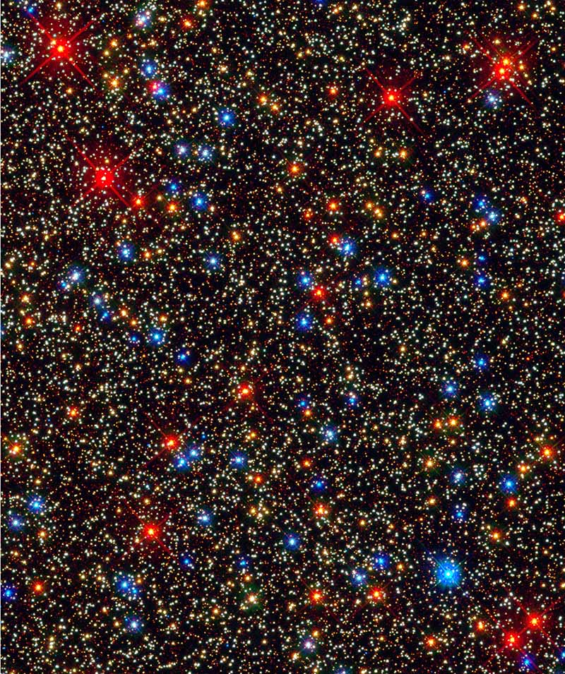 A black background is speckled by a seemingly infinite number of bright spots, with smaller spots being a pale-yellow color with some brighter and larger spots that are orange, bright red, and blue.