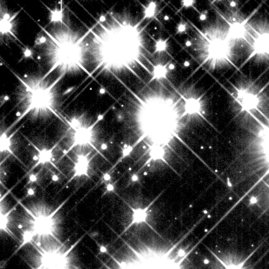 This field of stars is filled with white blobs with fuzzy edges, each a star in the globular cluster called M4. Each star has diffraction spikes that look like a large white X with the star at the center. There are about a dozen large stars that dominate the frame, with smaller ones filling in some of the gaps. A few are cut off by the edge of the image. 