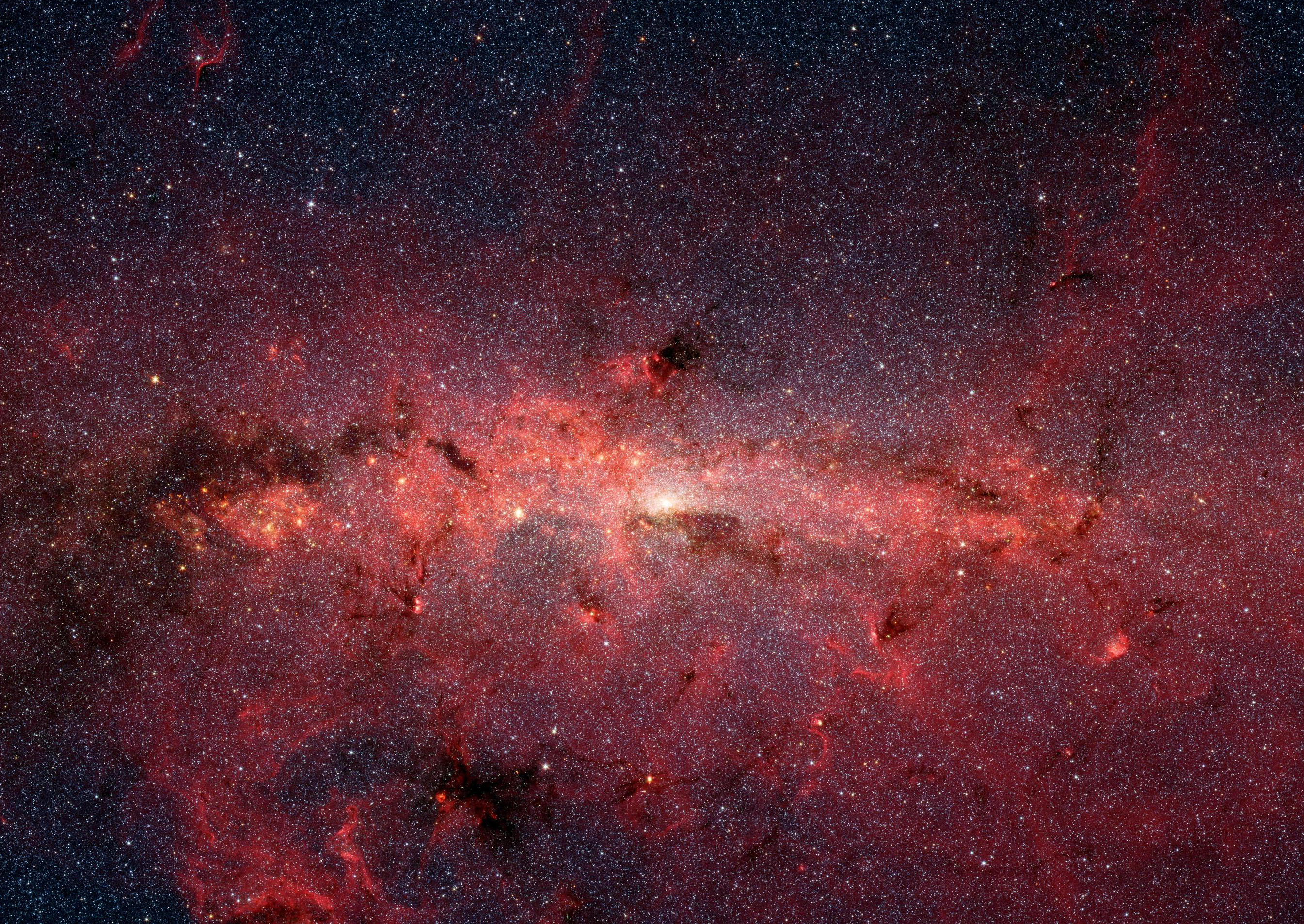 This image of the central region of the Milky Way shows a bright white glow at the center. An irregular line of light pink clouds stretches across the center of the image. Clouds in darker shades of pink and red spread out from that line, with more appearing at the bottom of the image than the top. Seemingly overlaid on these pink clouds are several dark maroon patches of different sizes. The background is studded with tiny white dots which are just some of the stars found in our galaxy. 