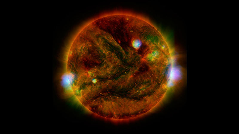 The Sun appears as a red-hued marble with black branching strips and few bright dots in white with small haloes of pale blue and electric green. All around the edge of the Sun is a glow, appearing like short fur in orange, yellow, and two bright spots of white on the right and left sides.