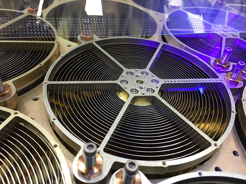 Taking up the center of the photo is one of NICER’s X-ray concentrators which is formed from concentric cylindrical mirrors that look like a vinyl record with very deep grooves. On top of the concentrator is a metal structure with a hexagonal plate in the center that has thin bars extending from each of the six corners, looking like a spiderweb. This one concentrator is surrounded by five others, which are only partially seen. Along the top quarter of the image is a clear window that reflects rectangular light and has a series of small holds in a grid across the bottom edge. Some parts of the central concentrator and the one to the upper right of it are bathed in purple light.
