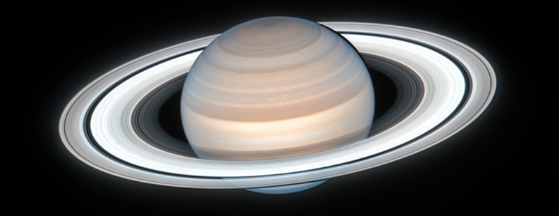 The ringed planet Saturn is seen on a stark black background. The round planet has bands of pale yellow, tan, and brown that appear as stripes across its surface, with the brightest and broadest band around the center of the planet and varying sizes of bands moving up to the top. Encircling the planet is a system of rings which are seen in front of the planet with part of the rings hidden behind the back. The rings also have bands of color ranging from pale blue to white and dusty tan with a few black areas showing gaps between the rings.