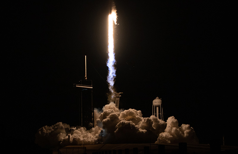 Against a dark background, a rocket launches. Visible is a nest of clouds on the ground with a bright plume of fire rising up to the top of the image. A structure is silhouetted to the left of the plume, and a water tower is visible just to the right.