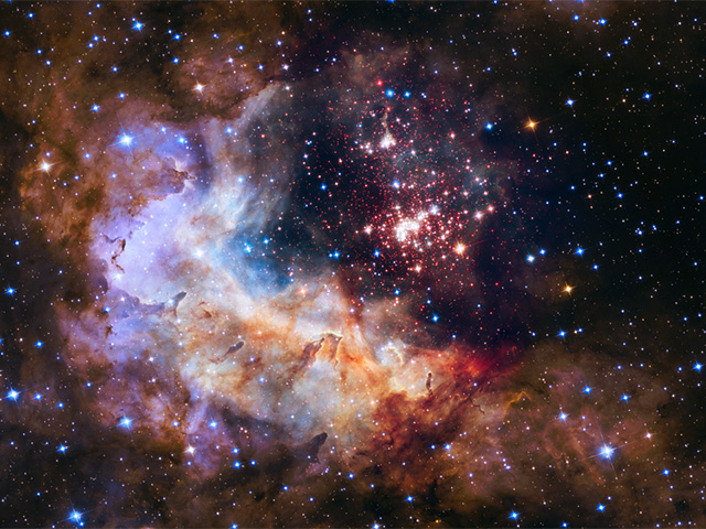 This image shows a dense cluster of stars just offset to the right from center. The stars in the cluster are predominantly white tinted with red. Across the entire image are additional stars with both red-tinted and blue-tinted stars. There is a pillowy nebula taking up the left side and bottom of the image. Within the nebula, several pillars of dark, dense gas are being shaped by the energetic light and strong stellar winds from the brilliant cluster of thousands of stars.