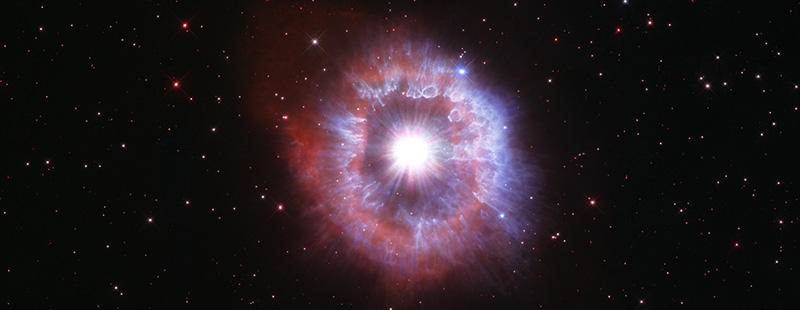 On a background dotted with small stars, AG Carinae appears as a giant eye in the sky. At the center is a bright white ball of light, and surrounding it is a ring of electric blue light and pale red light.